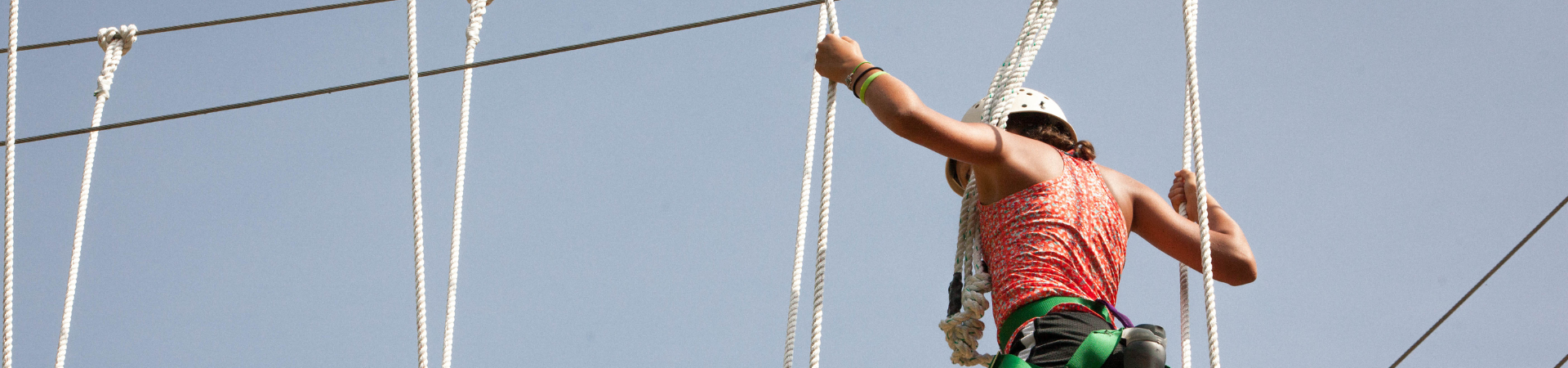  Girl in pink tank top and white helmet walking across a high ropes course 